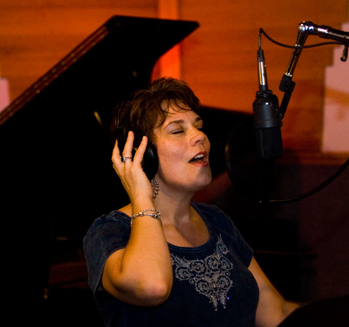 Jill Hatter singing into a studio microphone with a grand piano behind her